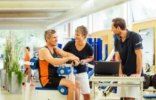 Ein Mann sitzt auf einem Fitnessgerät während zwei Sportlehrer bei ihm eine computergestützte Muskelfunktionsanalyse durchführen