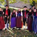 Gruppe von sieben Personen in traditioneller schwarzwälder Tracht, bestehend aus roten, blauen und lila Röcken, weißen Hemden und schwarzen Westen. Sie stehen auf einem mit Laub bedeckten Boden.