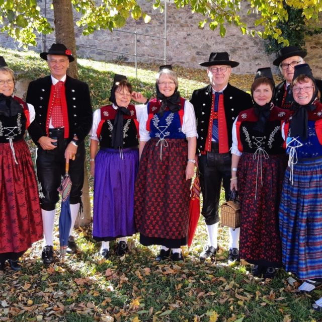 Gruppe von sieben Personen in traditioneller schwarzwälder Tracht, bestehend aus roten, blauen und lila Röcken, weißen Hemden und schwarzen Westen. Sie stehen auf einem mit Laub bedeckten Boden.