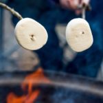 Zwei Personen halten jeweils einen Stock mit Marshmallows drauf über ein Feuer