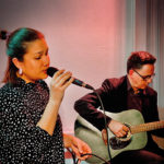 Eine Frau im schwarzen Kleid mit weißen Punkten singt in ein Mikrofon und ein Mann im schwarzen Anzug begleitet sie auf einer Gitarre.